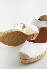 Valged espadrillid kõrged kontsad, mille pealmine osa on valmistatud kangast, punutud jute vaheosa ja kummist välistald. Omab pruuni kangast varbakatet ja pahkluu rihma.