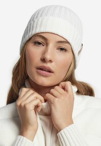 Hvid strikket hue med ribbet tekstur og et lille accentmærke. Modellen bærer en matchende hvid sweater med høj krave, hænderne foldet ved halsen.