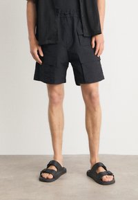 Shorts cargo noirs avec plusieurs poches, taille à cordon de serrage, et coupe décontractée, associés à des sandales noires à large bride et détail de boucle.