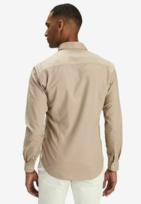 Beige långärmad skjorta med krage, knappade manschetter och en slät textur, med en ryggsyd och subtila sidosprund.