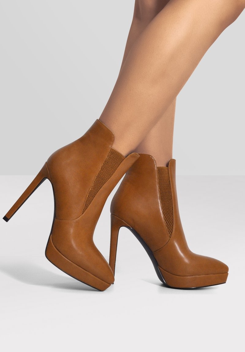 Jambes portant des bottines marron à talons hauts avec semelles compensées, panneaux latéraux élastiques et bouts pointus sur un fond gris uni.