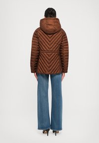 Kvinne med mørkt hår i en knute, iført brun quiltet hettejakke, blå vide jeans og sorte hæler, som står med ryggen til mot hvit bakgrunn.