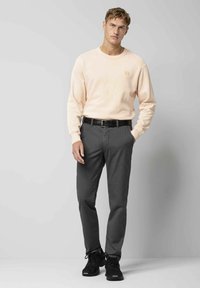 Helles rosa Sweatshirt mit langen Ärmeln und kleinem Logo, kombiniert mit dunkelgrauen Hosen. Schwarze Turnschuhe runden das Outfit ab.