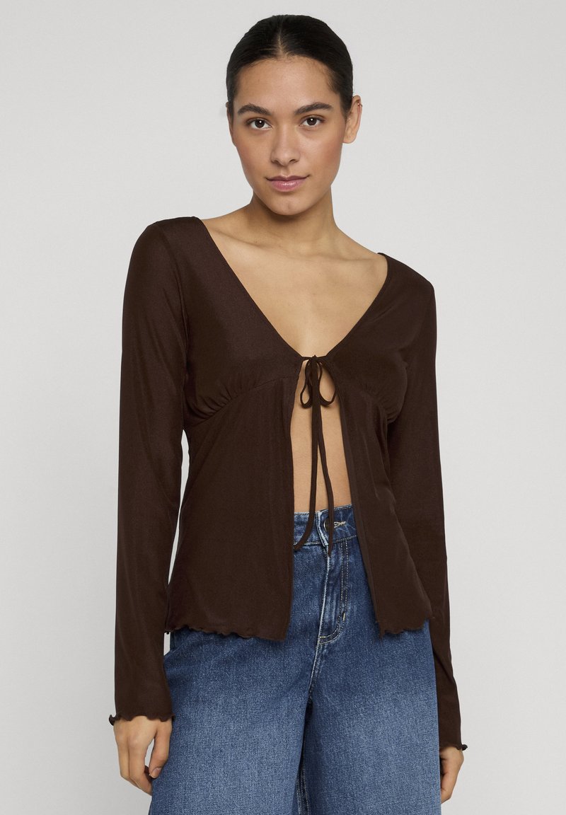 Femme portant un haut marron foncé à manches longues avec un profond décolleté en V et un nœud devant, associé à un jean bleu taille haute sur un fond uni.