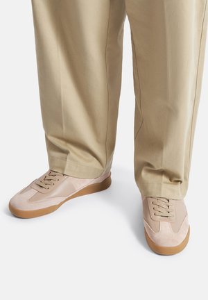 Bēša krāsas sporta apavi ar gumijas zolēm, valkāti ar bēša brīva piegriezuma biksēm uz balta fona, rādot ikdienišķu, neitrālas krāsas apģērba detaļu.