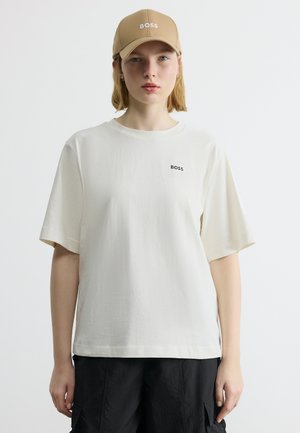 Personne portant une casquette BOSS beige, un t-shirt BOSS blanc et un pantalon cargo noir, debout devant un fond clair uni.