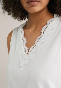 Fehér ujjatlan felső, V-nyakkivágással, bonyolult csipke díszítéssel és fogazott szélekkel, amely a nyakrészen texturált részleteket mutat.