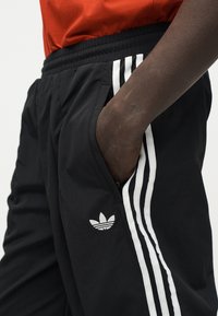 Άτομο με σκουρόχρωμο δέρμα που φοράει μαύρο παντελόνι Adidas με λευκές ρίγες, με το χέρι στην τσέπη και ένα κόκκινο τοπ ορατό στη μέση.