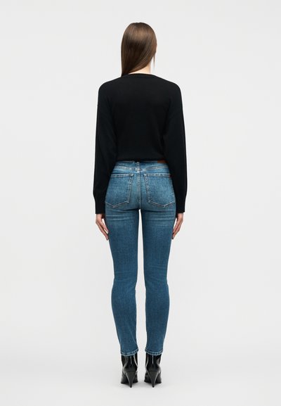 Γυναίκα με καστανά ίσια μαλλιά, φοράει μαύρο πουλόβερ μακριάς μανίκας, στενά μπλε τζιν και μαύρα μπότες με τακούνι μέχρι τον αστράγαλο, στέκεται με την πλάτη προς το μέρος μας.