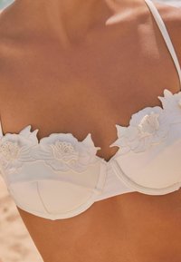 Vit bikinitopp med blommönster broderat längs halsringen, med justerbara remmar och en strukturerad kupdesign.