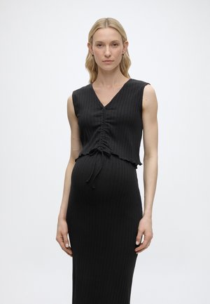 Femme enceinte aux longs cheveux blonds, portant une robe noire côtelée sans manches avec un devant froncé et une taille resserrée par un cordon, debout devant un fond uni.