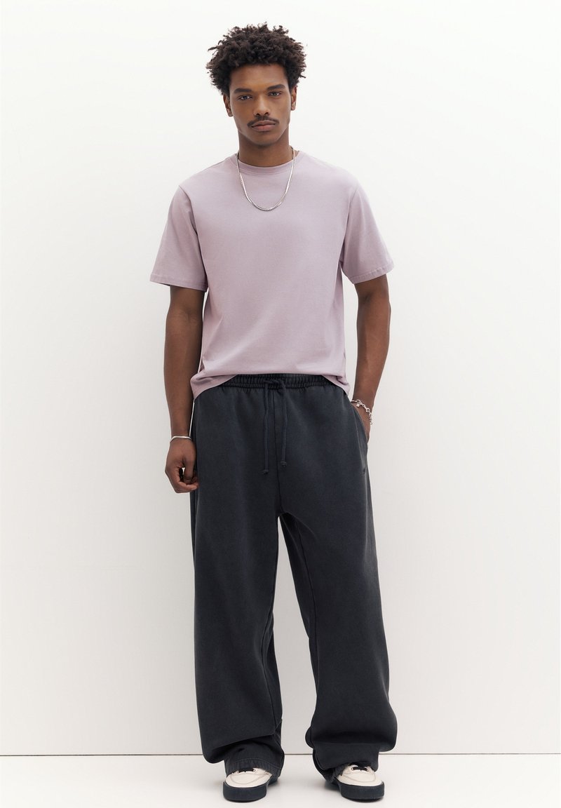Jeune homme aux cheveux bouclés portant un t-shirt violet clair, un pantalon noir ample, des baskets blanches, une chaîne en argent et des bracelets, debout contre un mur blanc.
