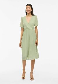 Femme portant une robe midi plissée vert clair avec des manches courtes à volants et des sandales à talons hauts dorées à lanières, se tenant devant un fond blanc.