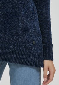 Pulover tricotat de culoare albastru marin, cu o suprafață texturată, manșete cu nervuri și un mic accent metalic pe marginea laterală. Material moale și voluminos.