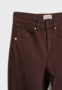 Jeans en denim brun foncé avec une taille haute, des accessoires en cuivre, deux poches avant et une étiquette de marque sur la ceinture.