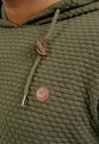 Sudadera verde oliva con tela texturizada, que presenta un parche de cuero redondo y un cordón trenzado con acentos metálicos.