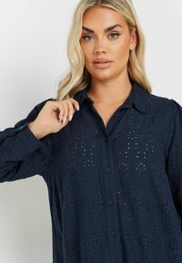 Blusa blu navy con un intricato motivo a occhio, chiusura a bottoni davanti e collo con risvolto. Tessuto testurizzato e maniche lunghe a sbuffo.