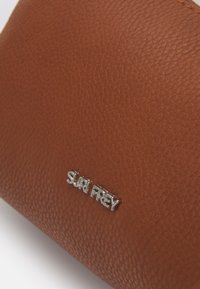 Hnědý texturovaný kožený povrch s připevněným stříbrným kovovým logem značky "SURI FREY".