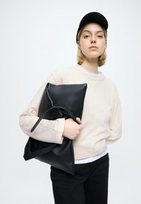 Pochette en cuir noir avec un design matelassé et une bandoulière amovible, tenue contre un pull beige clair, avec une chemise blanche en dessous.