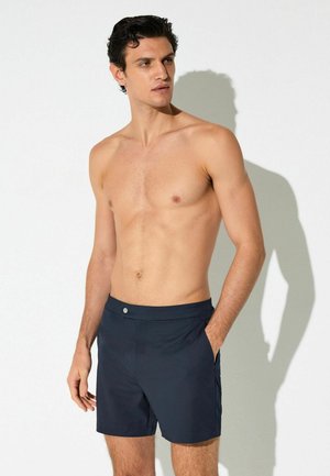 Skjorteløs mand med mørkt krøllet hår iført marineblå badeshorts, stående med den ene hånd i lommen, kigger til siden mod en hvid baggrund.