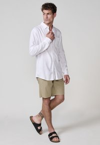 Cămașă albă cu mânecă lungă și nasturi, prevăzută cu un buzunar pe pieptul stâng, combinată cu şorturi bej și sandale negre, evidențiind un fit relaxat și un stil casual.