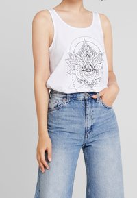 Débardeur blanc avec un motif de lotus noir imprimé, associé à un jean en denim léger bleu clair taille haute. Tissu lisse, coupe décontractée, sans embellissements visibles.