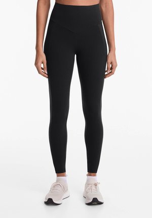 Zwarte high-waisted leggings van gladde stof, met accent naden en taps toelopende benen, gecombineerd met lichtgekleurde sportschoenen.