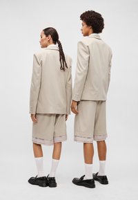 Blazer e calções bege claro, de corte ajustado, tecido liso, com bainha enfrentada nos calções, acompanhados de meias brancas e sapatos pretos.