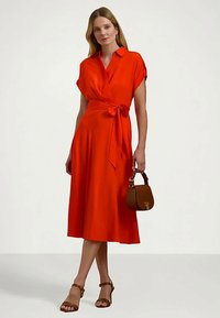 Γυναίκα σε φωτεινό κόκκινο φόρεμα με τύλιγμα, κρατώντας μια καφέ τσάντα, φορώντας καφέ πέδιλα με τακούνι, που στέκεται μπροστά σε απλό ανοιχτό φόντο.