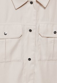 Krämfärgad skjorta med två fickor på bröstet, svarta metallknappar med tryck, och en spetsig krage, som visar tygtextur och sömnadsdetaljer.