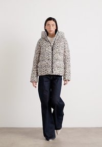 Ein Model läuft selbstbewusst in einer stilvollen Leopardmuster-Daunenjacke und dunklen, weit geschnittenen Jeans vor einem neutralen Hintergrund.