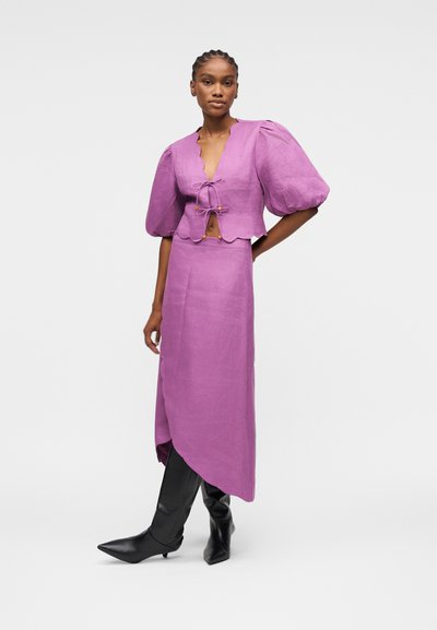 Femme portant un chemisier court violet à manches ballon avec des liens à l'avant et une jupe midi asymétrique assortie, associée à des bottes noires pointues.