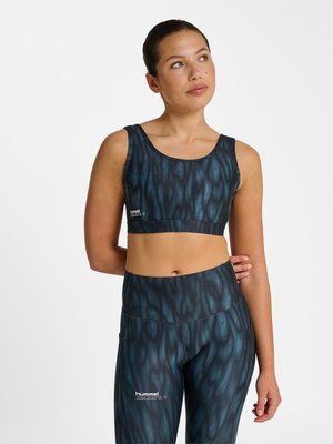 Femme portant un soutien-gorge de sport et un legging noir et bleu à motifs, debout avec un bras derrière le dos, regardant en haut et à droite.