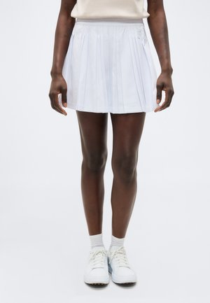 Person wearing a white pleated tennis skirt, white socks, and white athletic shoes, standing against a plain background.