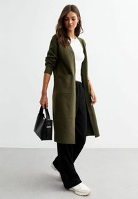 Cardigan lungo color verde oliva con polsini a coste, apertura frontale e due tasche, abbinato a una maglietta bianca e pantaloni neri, mentre tiene una borsa nera.