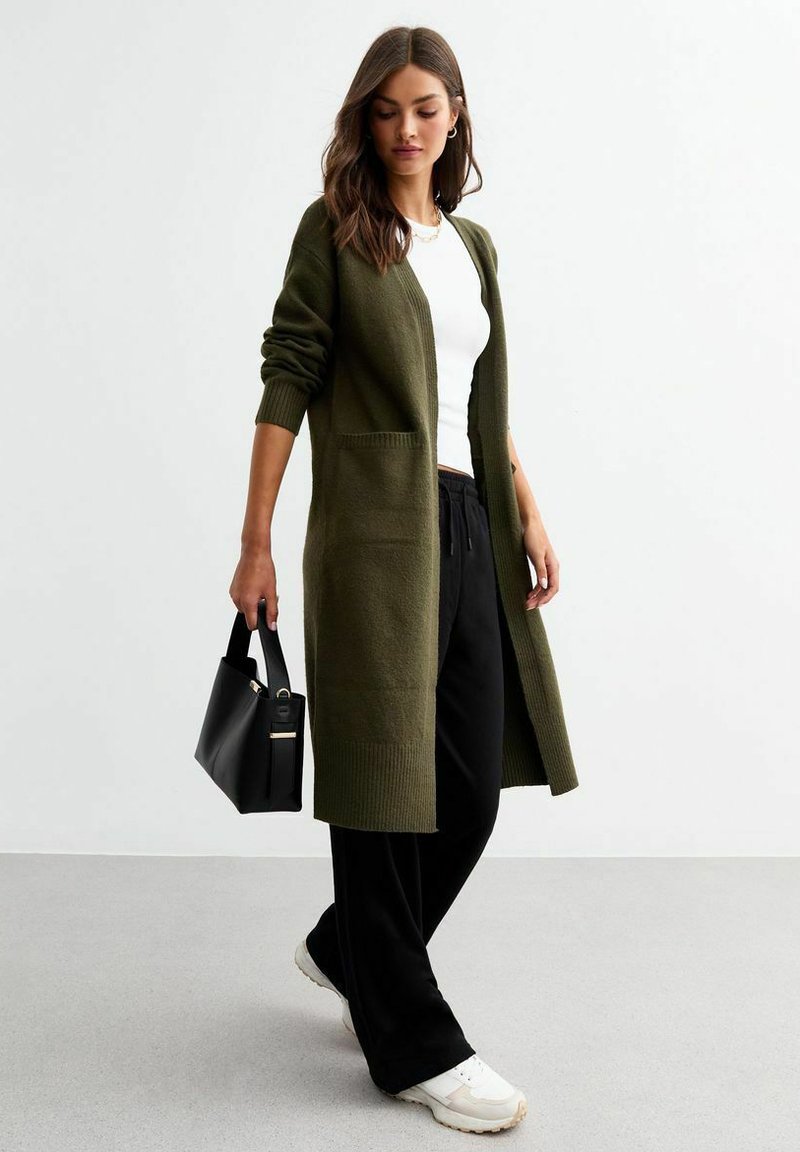 Cardigan lungo color verde oliva con polsini a coste, apertura frontale e due tasche, abbinato a una maglietta bianca e pantaloni neri, mentre tiene una borsa nera.