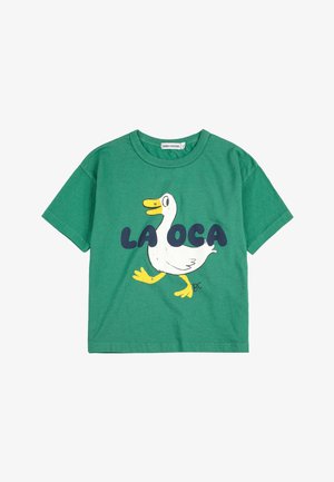 Grünes Baumwoll-T-Shirt mit einem weißen Entenmotiv, das einen gelben Schnabel und Füße hat, begleitet von dem Text "LA OCA" in fetten dunkelblauen Buchstaben.