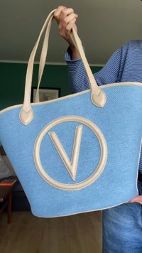 Zila auduma iepirkumu soma ar bežām ādas rokturiem un apaļu logotipu. Auduma virsma ir gluda, un soma ir plašā dizainā.