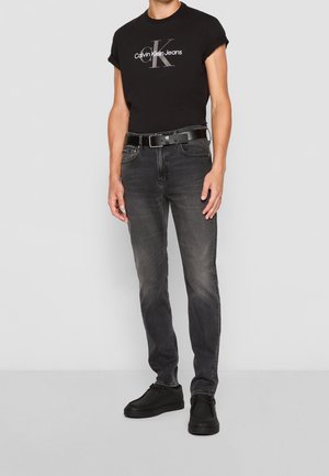 Homme portant un t-shirt noir Calvin Klein Jeans, un jean noir délavé avec une ceinture noire et des chaussures noires, debout devant un fond clair uni.