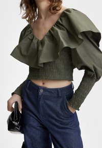 Top corto arricciato verde oliva con design arricciato; abbinato a pantaloni in denim blu scuro a vita alta. Una borsa nera completa il look.