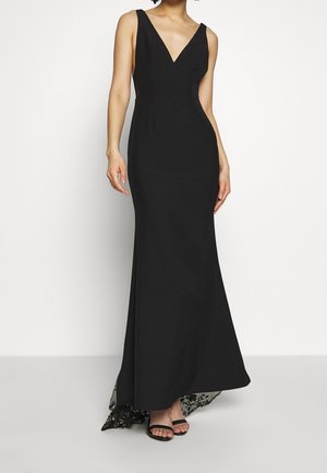 Nainen pukeutunut mustaan, hihattomaan iltapukuun, jossa on syvä V-pääntie ja pitsikoristeltu helma, yhdistettynä mustiin avokkaisiin.