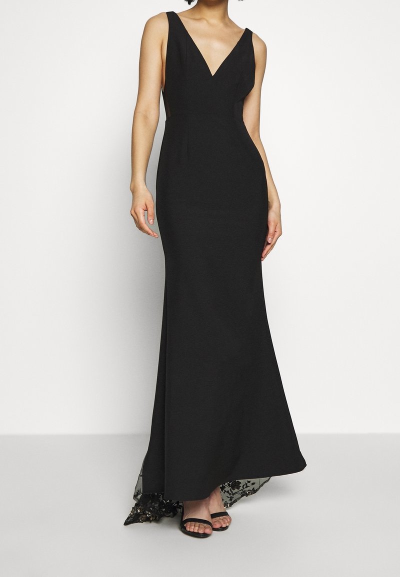 Femme portant une robe de soirée noire sans manches avec un décolleté en V profond et un ourlet orné de dentelle, associée à des escarpins noirs à bout ouvert.