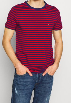 Man wearing a red and navy blue horizontal striped t-shirt with short sleeves and blue distressed jeans, hands in pockets.