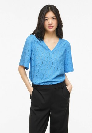 Žena nosi textúrovaný modrý top s krátkym rukávom a výstrihom do V a čierne nohavice s vysokým pásom, s rukami v vreckách, stojaca pred bielym pozadím.