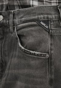 Γκρι τζιν από ντένιμ με φθαρμένη τσέπη, μαύρη κεντημένη ετικέτα που αναγράφει «REPLAY» και ασημί μεταλλικά στοιχεία στη μέση.