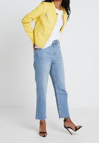 Kvinna iklädd en gul kort jacka, vit grafisk t-shirt, ljusblå jeans med fransar och svarta högklackade sandaler, stående mot vit bakgrund.
