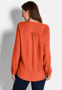 Langärmlige Bluse aus orangefarbigem Stoff mit plissiertem Rückendesign, abgerundetem Saum und geknöpften Manschetten. Glatte Textur und lässige Passform.