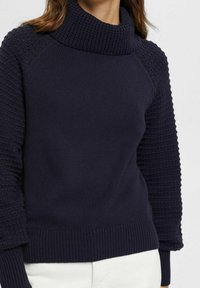 Pull en maille bleu marine avec un col cheminée haut. Comprend des manches texturées et un ourlet côtelé. Conception légère et ajustée.