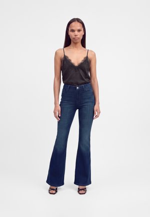 Woman with long dark hair wears black lace-trimmed camisole, dark blue flared jeans, and black open-toe heels, standing against plain background.