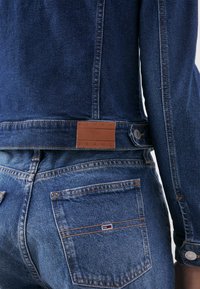 Tmavě modrá džínsová bunda s hnědou koženou nášivkou s nápisem "TOMMY JEANS," s oranžovým prošíváním a detailem kapsy.